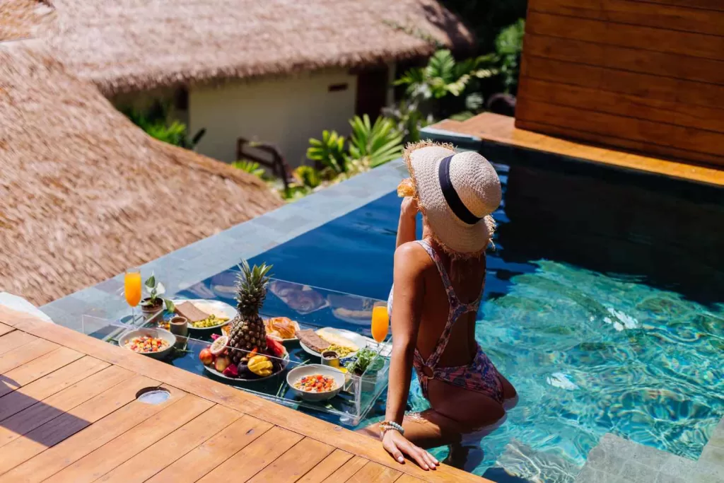 Beautiful caucasian tanned woman in bikini and straw hat with floating breakfast at amazing luxury bali style villa at sunny day by swimming pool