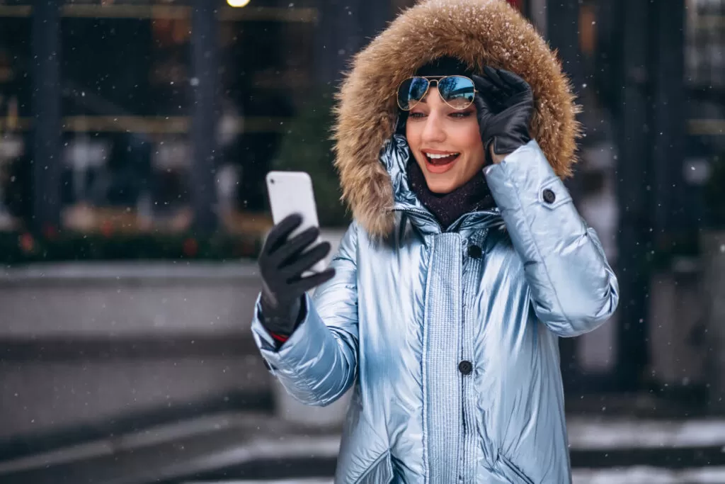 A girl in winter gear and sunglass looking at her phone with surprise face.